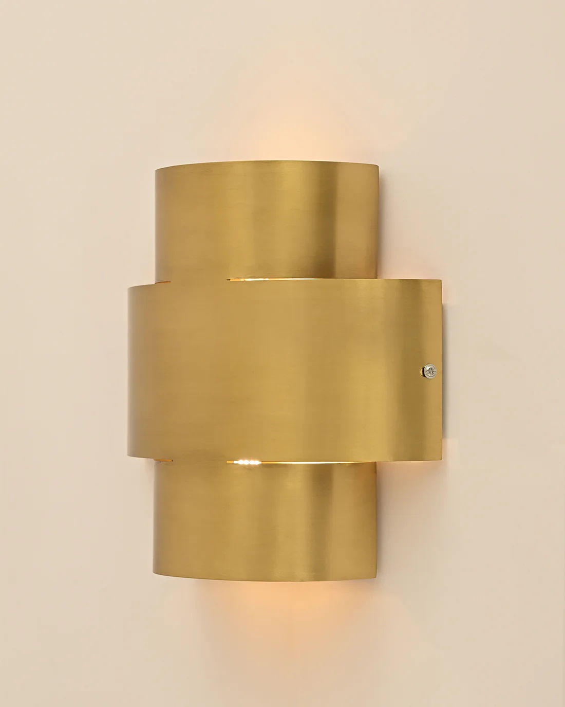 The Aura Wall Sconce by LMNOH features a sleek, brushed brass finish with overlapping cylindrical shapes in a cross pattern. It complements contemporary interiors and emits a soft glow against neutral walls.