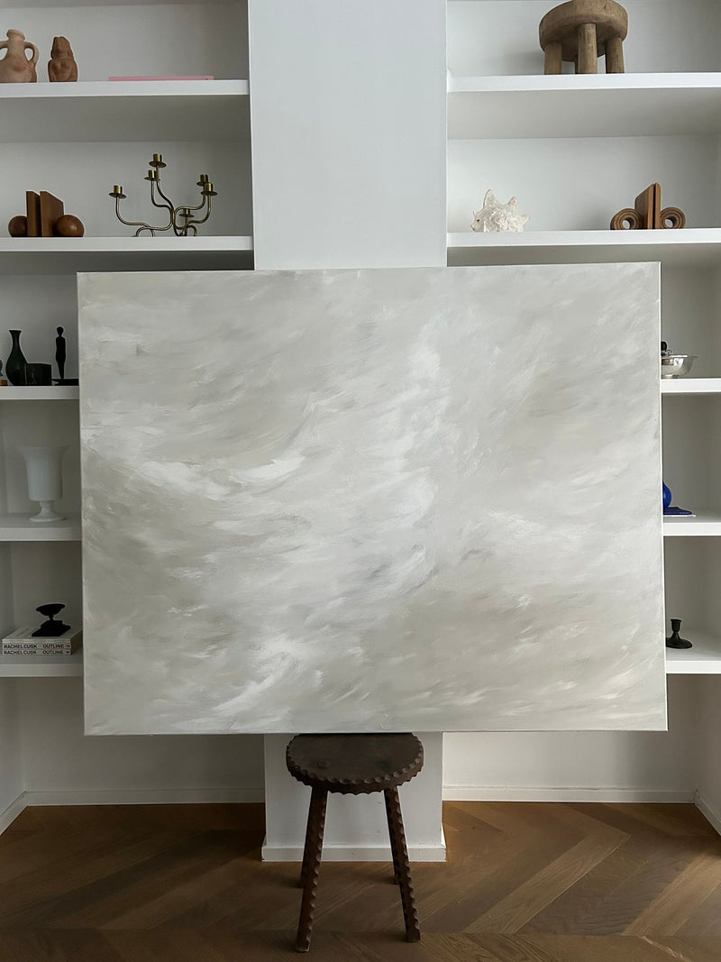 The "Whispering Breeze" painting by Mattias Storm, a large mixed media piece with light grey and white swirls, rests on a wooden stool. It complements white shelves decorated with sculptures, vases, and a candelabra above a classic herringbone floor.