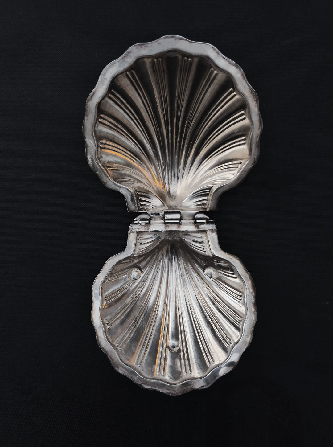 The Les Objoies Silver-plated Shell Box, resembling a clam shell, features hinged mechanics with ribbed and smooth textures. Its open metallic design shines against a dark backdrop, exuding a reflective charm perfect for home decor.