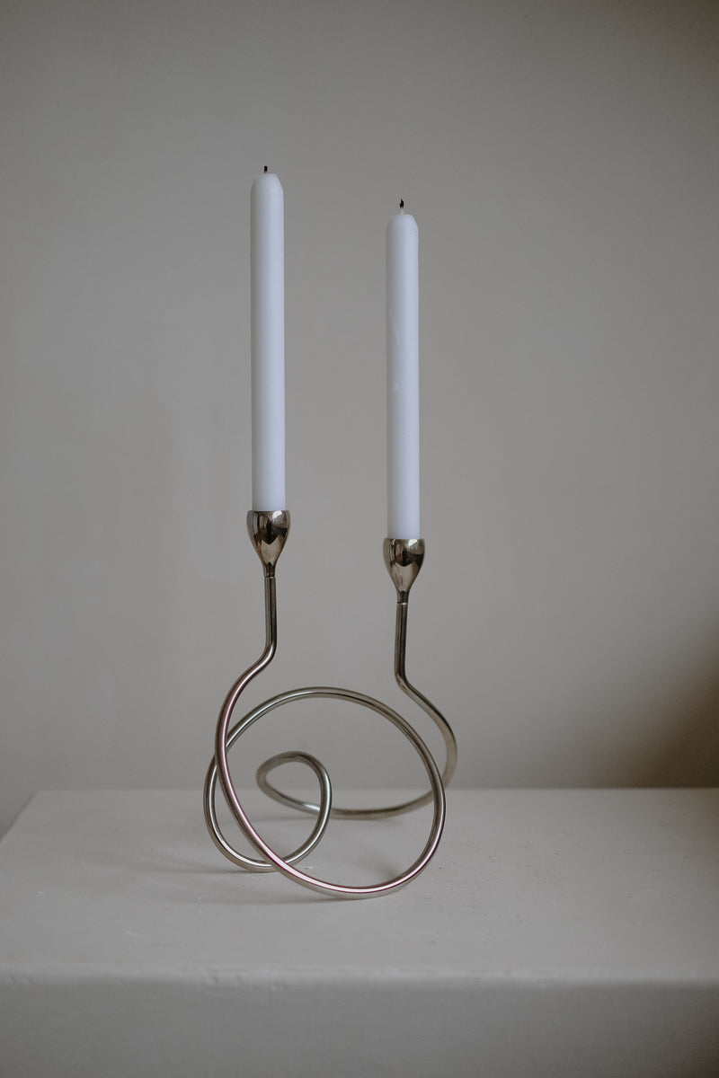 The septembre studios "Loop" Candle Holder showcases a minimalist chrome design with swirling loops and features two unlit white dinner candles, set against a neutral background.