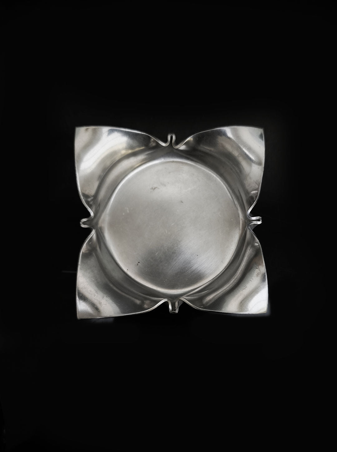 The Les Objoies Folded Ashtray features a polished silver finish with a round center and four petal-like corners, set against a black background, resembling folded brass.