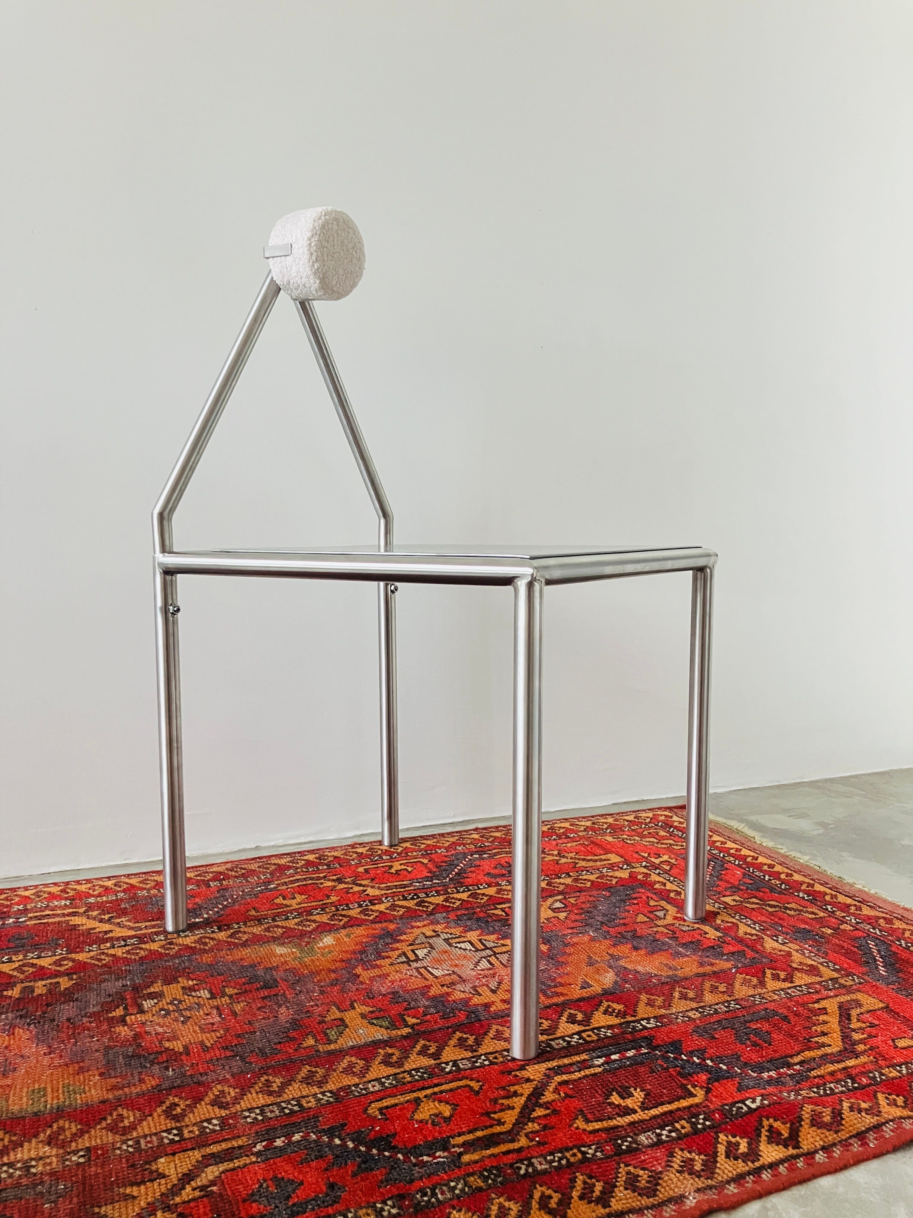 The 'H01' Stainless Steel Chair by HT, featuring an angled backrest and round white removable cushion headrest, sits on a red patterned rug against a plain white wall.