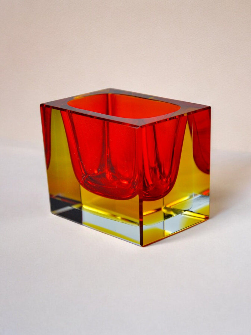 The Sommerso Ashtray by Seguso from Malamar Studio resembles Murano artistry with its clear glass cube, featuring a vibrant red and yellow inner design with striking curves and sharp angles. It rests on a plain, light surface casting subtle shadows.