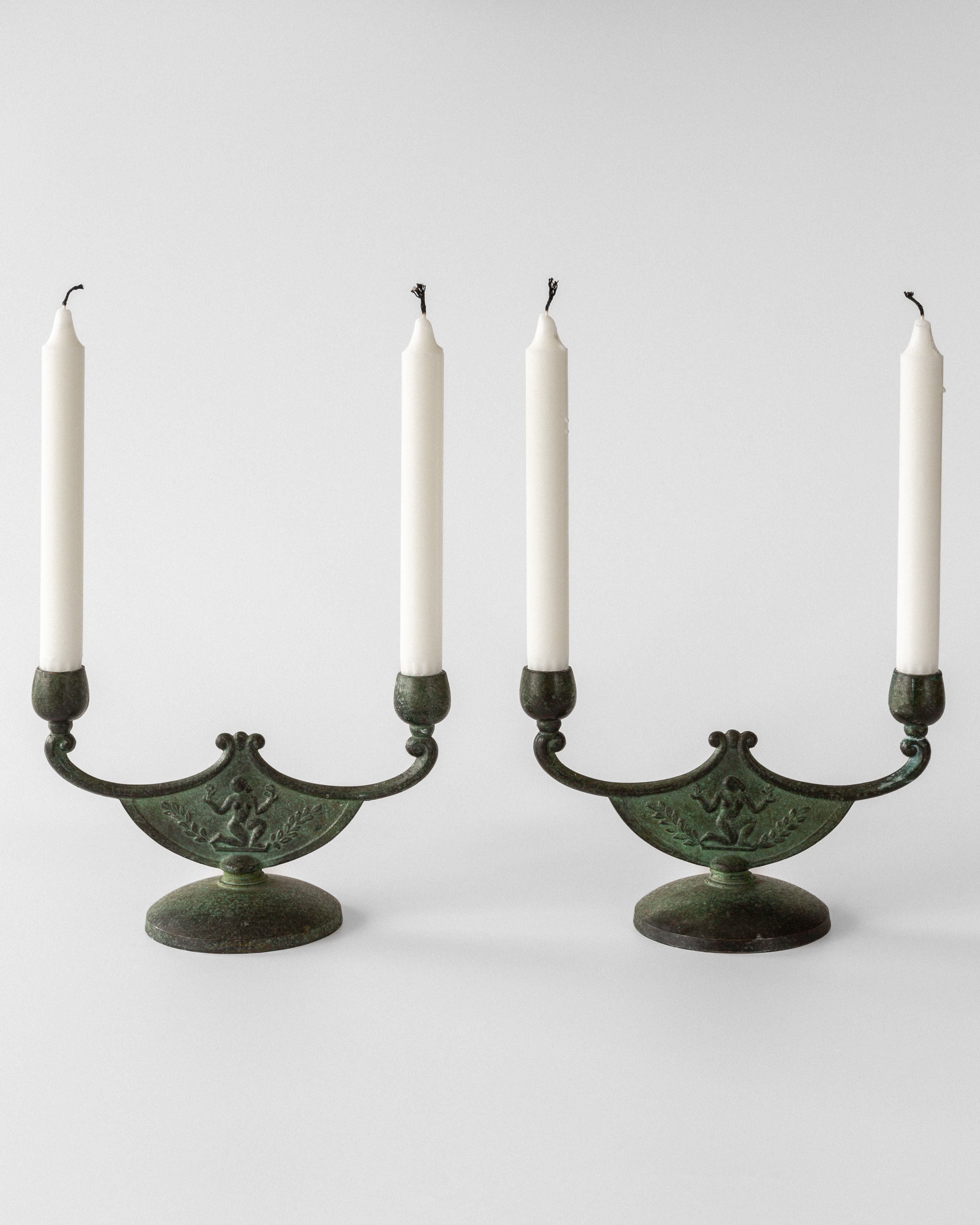 The Spigel Pair of Art Deco Bronze Candleholders exhibit exquisite Swedish metalwork with intricate designs and rounded bases, each holding a tall white candle, set against a plain white backdrop.