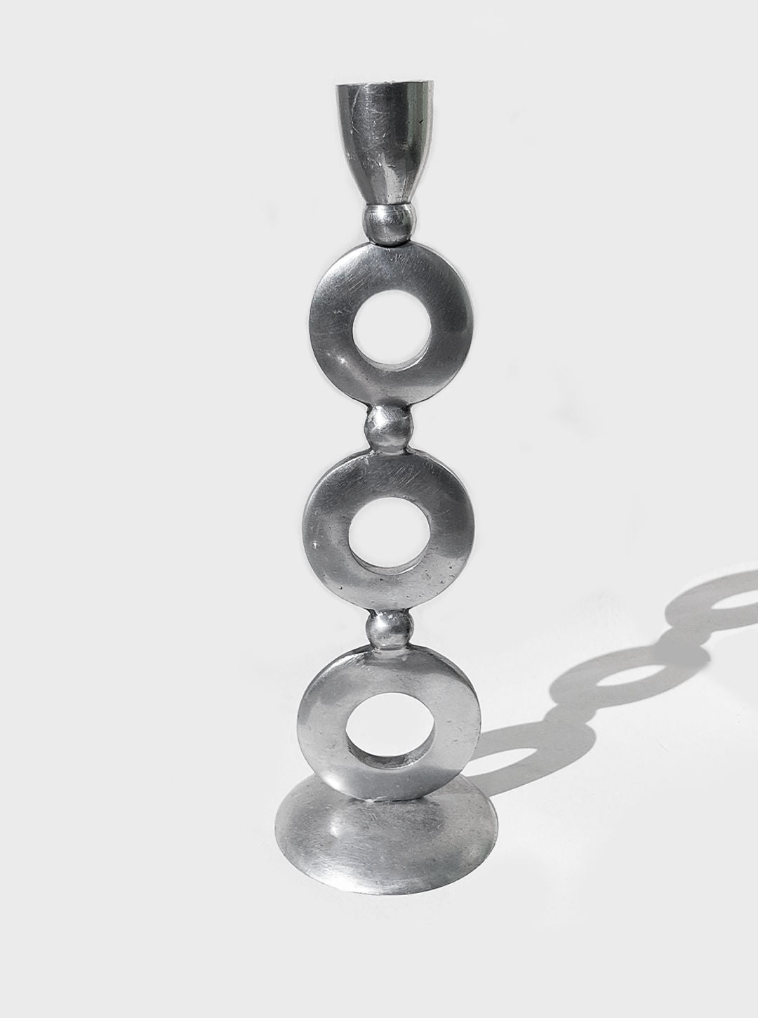 The Totem Candle Holder by Les Objoies is a sleek, modern metal candlestick holder with elegant silver plating. It features three circular discs stacked vertically, interspersed with small spheres. The round base supports a cylindrical holder for a candle at the top. Set against a white background that casts a shadow, this piece embodies contemporary design.