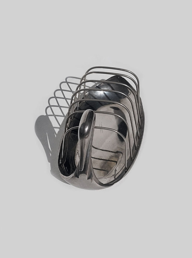 A collection of stainless steel kitchen utensils, including a whisk, measuring spoons, and a Les Objets brass swan toast rack, arranged neatly and casting a shadow on a light surface.