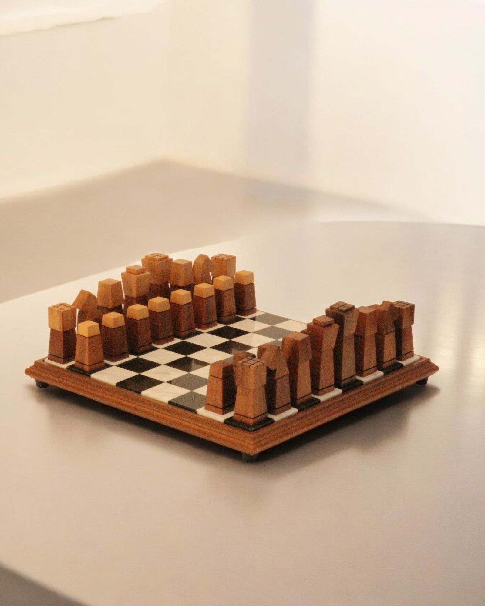 A Wooden and Marble Chessboard from Boga Avante Shop, boasting vintage design and abstract hand-carved chess pieces, is set up for a game. The chunky, stylized pieces feature geometric shapes, and the board is placed on a reflective surface in a minimalistic, warmly lit environment.