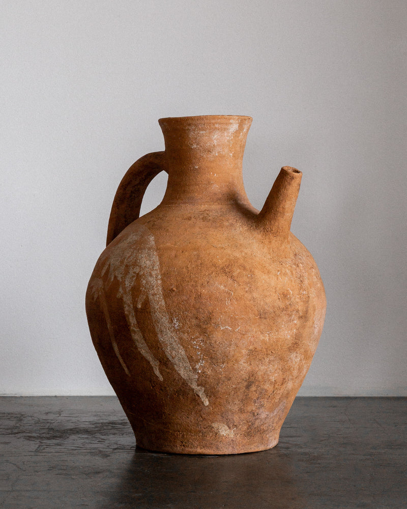 The Spigel Early 20th Century European Ceramic Pitcher, with its rustic design featuring a weathered finish, single handle, and short spout in earthy tones, rests on a dark surface against a plain, light gray background.