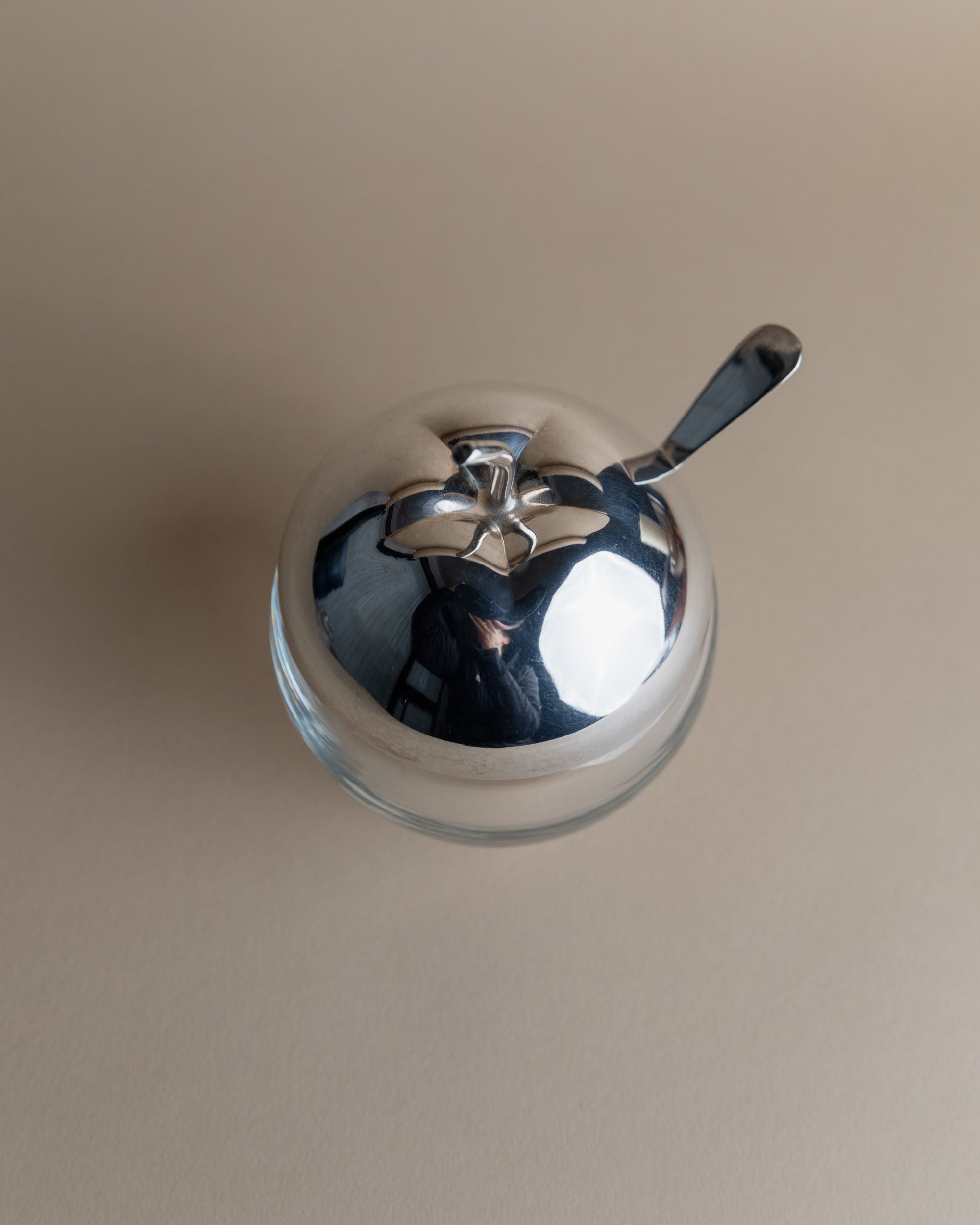 The Bottega Jacobs Silver & Glass Apple Jar, a reflective bowl with a silver-plated lid and spoon, sits on a light tan surface. The smooth lid mirrors its surroundings for an elegant touch.