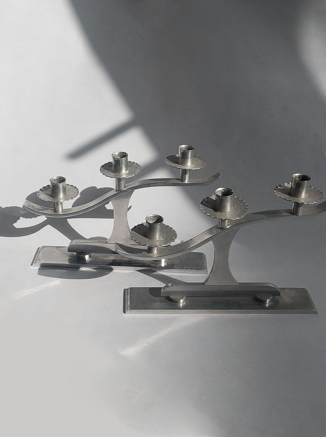 A photograph showcasing the Pair of Wavy Candle Holders by Les Objoies with a modern, metal design exuding Art Deco flair. Each holder boasts three candle spots and features a wavy pattern on rectangular bases. The candle holders cast intriguing shadows on the light background, adding dimension to the image.