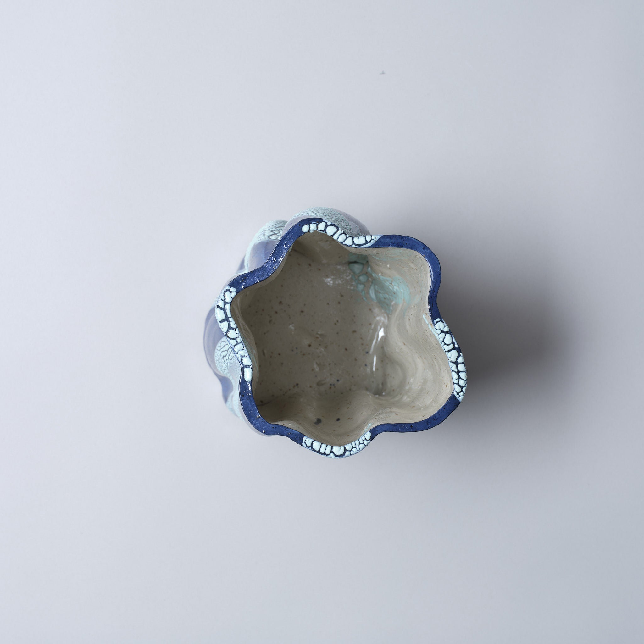 An overhead view reveals the Astrid Öhman Swirly Vase Blue and Light Blue, featuring a handmade wavy, flower-like rim. The exterior sports a blue hue with a white floral pattern, while the interior is speckled light gray. It rests on a simple, light gray background.