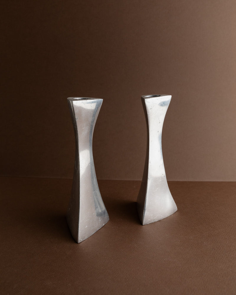 Two Bottega Jacobs "2 Aluminium Candleholders 80s" adorn a brown surface with a matching background. These candleholders feature distinctive twisted designs and a metallic finish, bringing a touch of retro-inspired ambiance to the space.