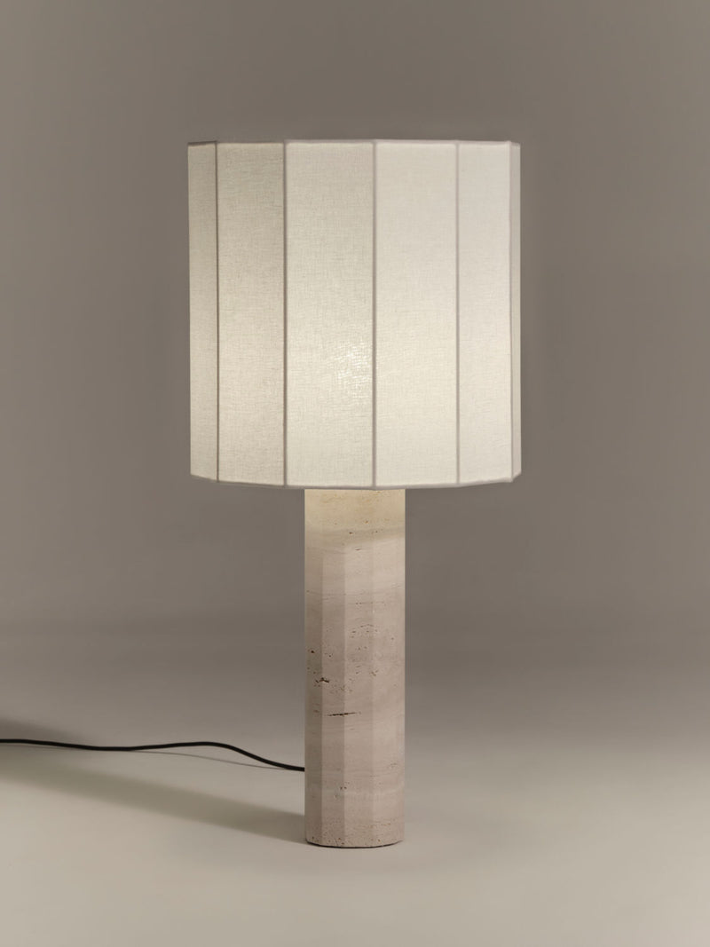 A Nassi Lamps Roma II Dodecagon White Travertine with a cylindrical base in a light stone texture reminiscent of white travertine, topped with a vertically paneled, white rectangular lampshade. The background is plain and grey, and a black power cord is visible on the left side.