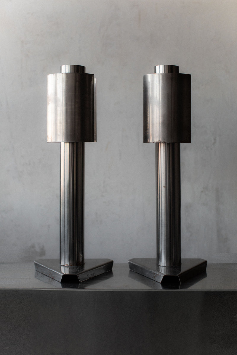 Two tall, cylindrical sculptures stand side by side on a flat surface. Each has a geometric, industrial design with a vertical orientation and a hexagonal base, exuding the 1960's Space Age aesthetic of Out For Lunch lamps against the plain gray background.