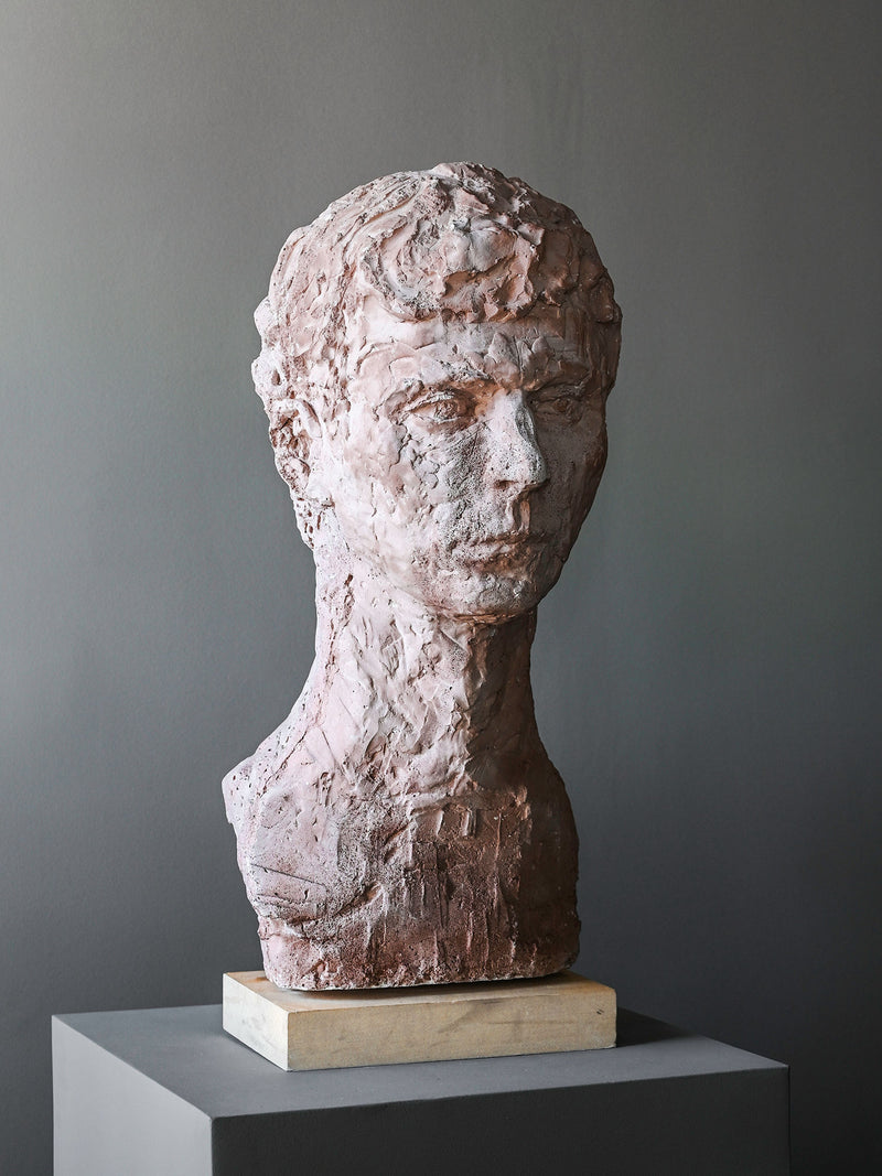 The “A Simple Man” Sculpture by Studio OSKLO is a textured, reddish-brown terracotta bust of a male figure with an elongated neck, rough chiseled details, and set on a light base against a plain gray backdrop.