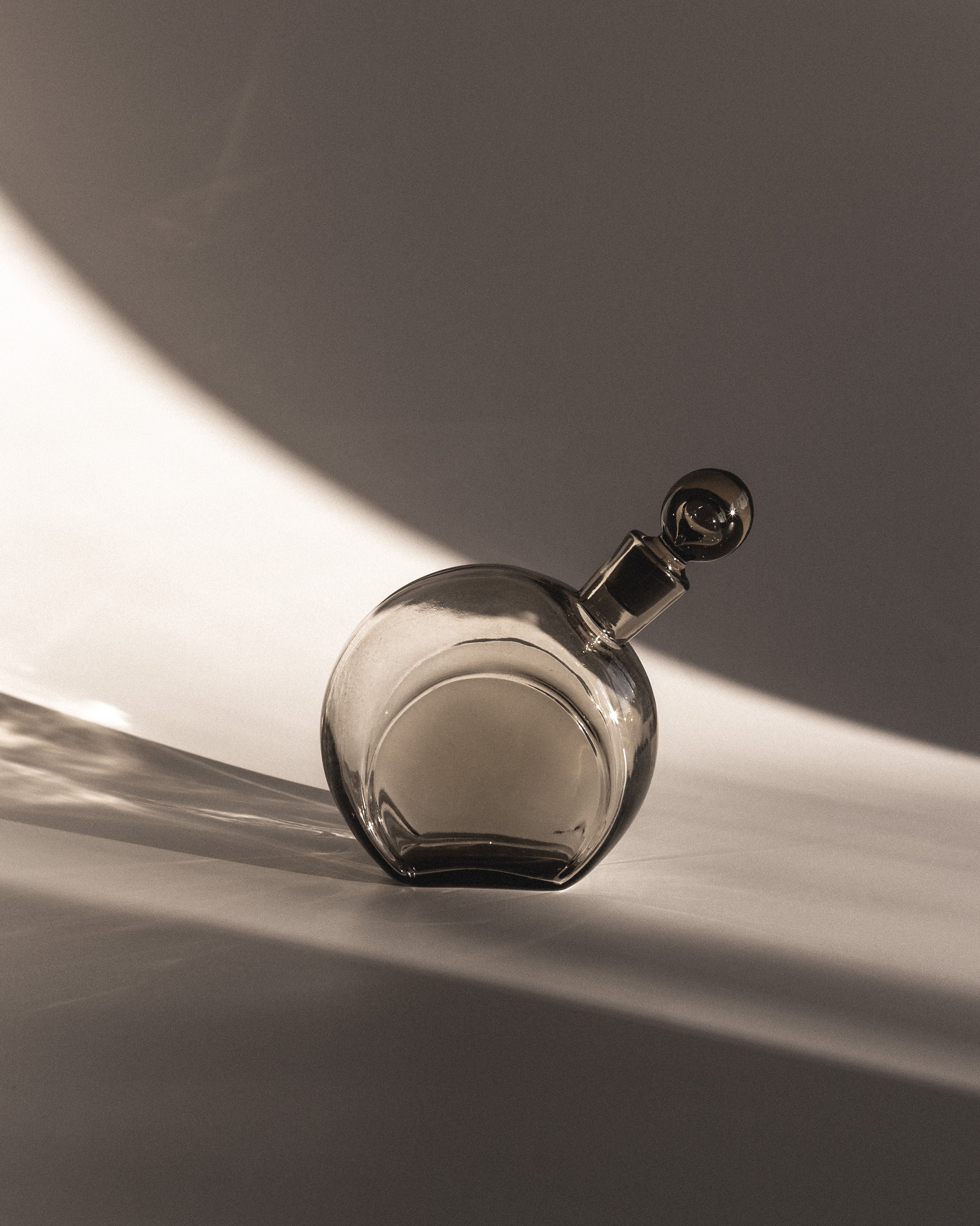 A round glass decanter by Harald Notini from the 1930s, featuring a smoky brown hue and a round stopper, rests on its side. Dramatic light and shadow patterns in the background evoke an elegant, minimalist aesthetic typical of Swedish modernism. Brand: Spigel.