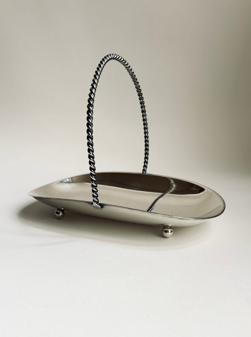 The Art Deco Silver Plated Basket by Flock Studio features a sleek, silver metallic design with a twisted handle and four small ball feet, exuding timeless vintage elegance reminiscent of an Art Deco piece, all beautifully presented against a plain light background.