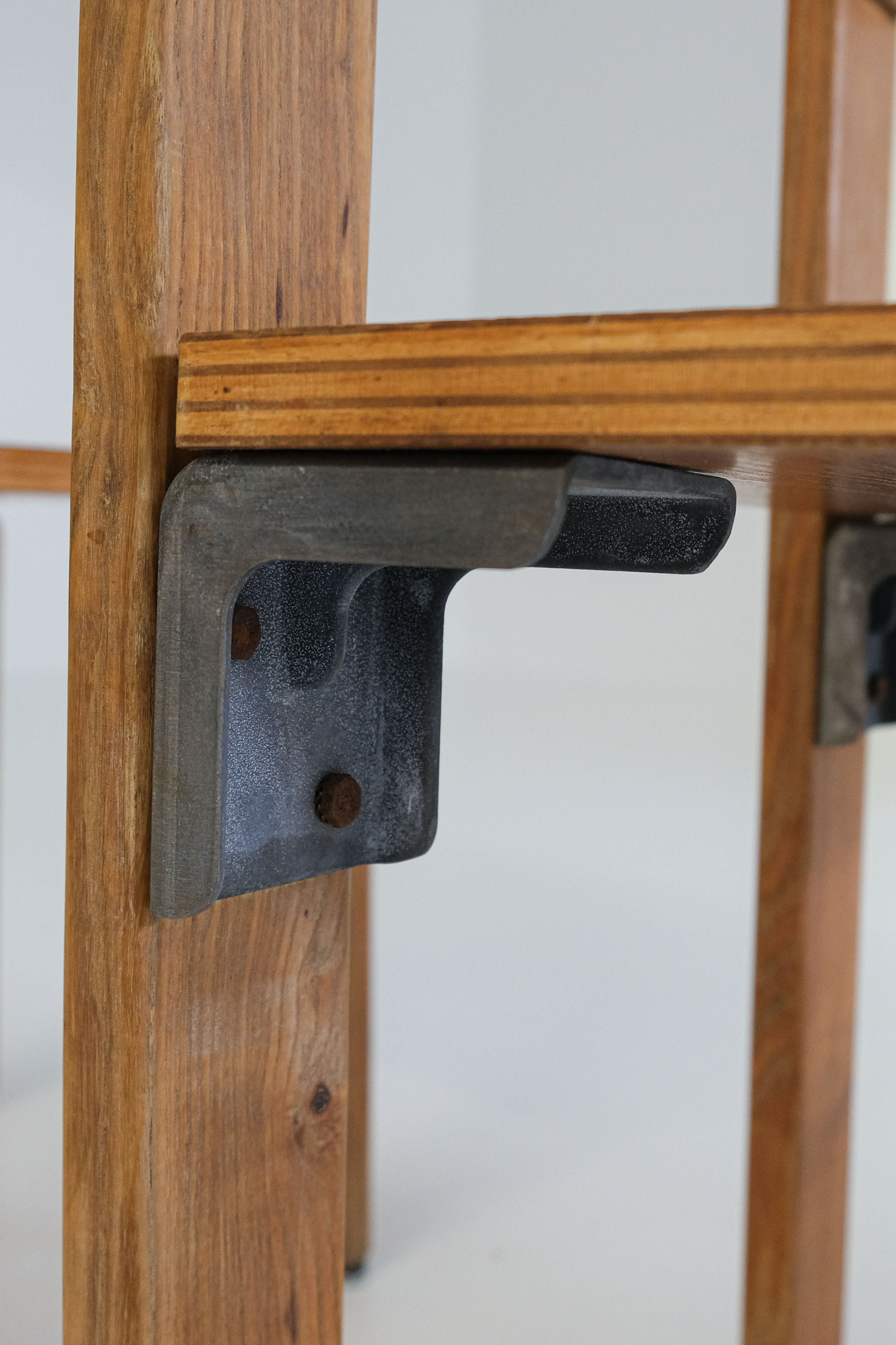 A vintage oak plywood shelf with a natural finish is secured by a side-attached cast aluminum bracket, showing slight rust for character against a plain white wall. Featured Product: "Les Carats" Dining Chairs by George Candilis and Anja Blomstedt 1970s, Brand: antibeige.