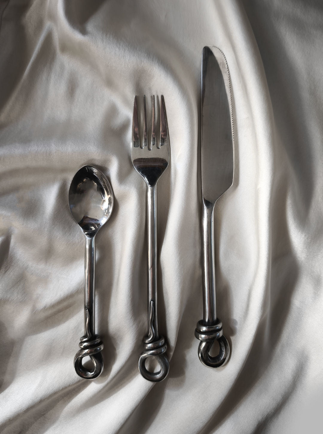 A Set of 3 twist cutlery by Les Objoies, featuring a spoon, fork, and knife with elegantly curved handles, is beautifully displayed on a smooth, silky fabric background, capturing the timeless charm of vintage design.