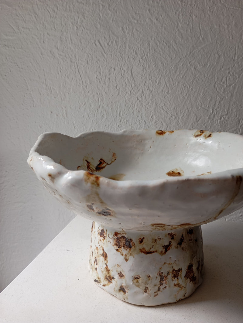 The Coupe 53 by Claire Pain is a handmade stoneware bowl with an irregular rim, a short wide pedestal base, and a white glaze with earthy brown spots—shown on a light surface against a textured white wall.