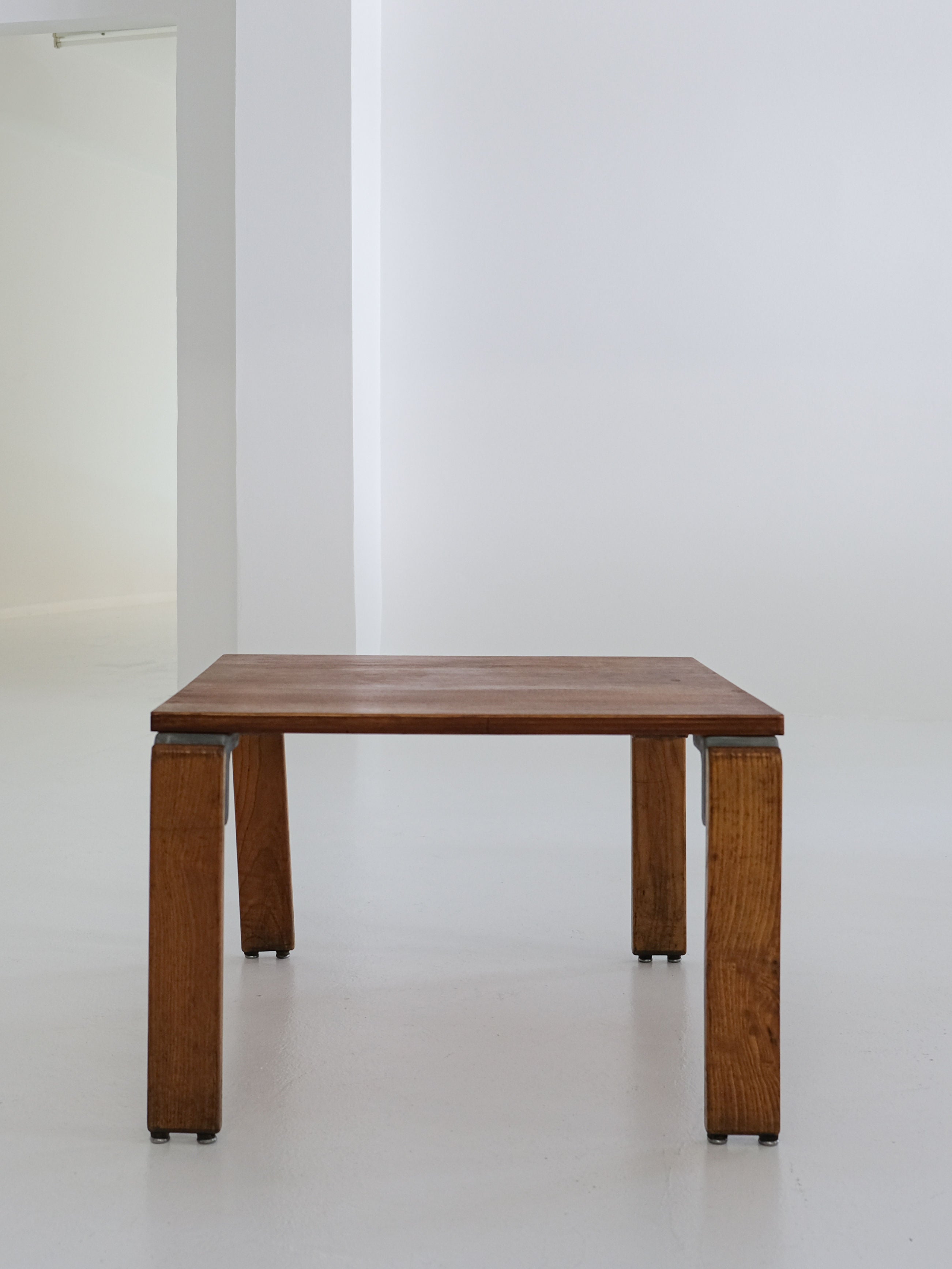 In a minimalistic white room with smooth walls and a bare floor, the antibeige Coffee Table by George Candilis and Anja Blomstedt from the 1970s stands. Its thick legs and natural oak plywood wood grain are highlighted by soft lighting, showcasing its vintage design.