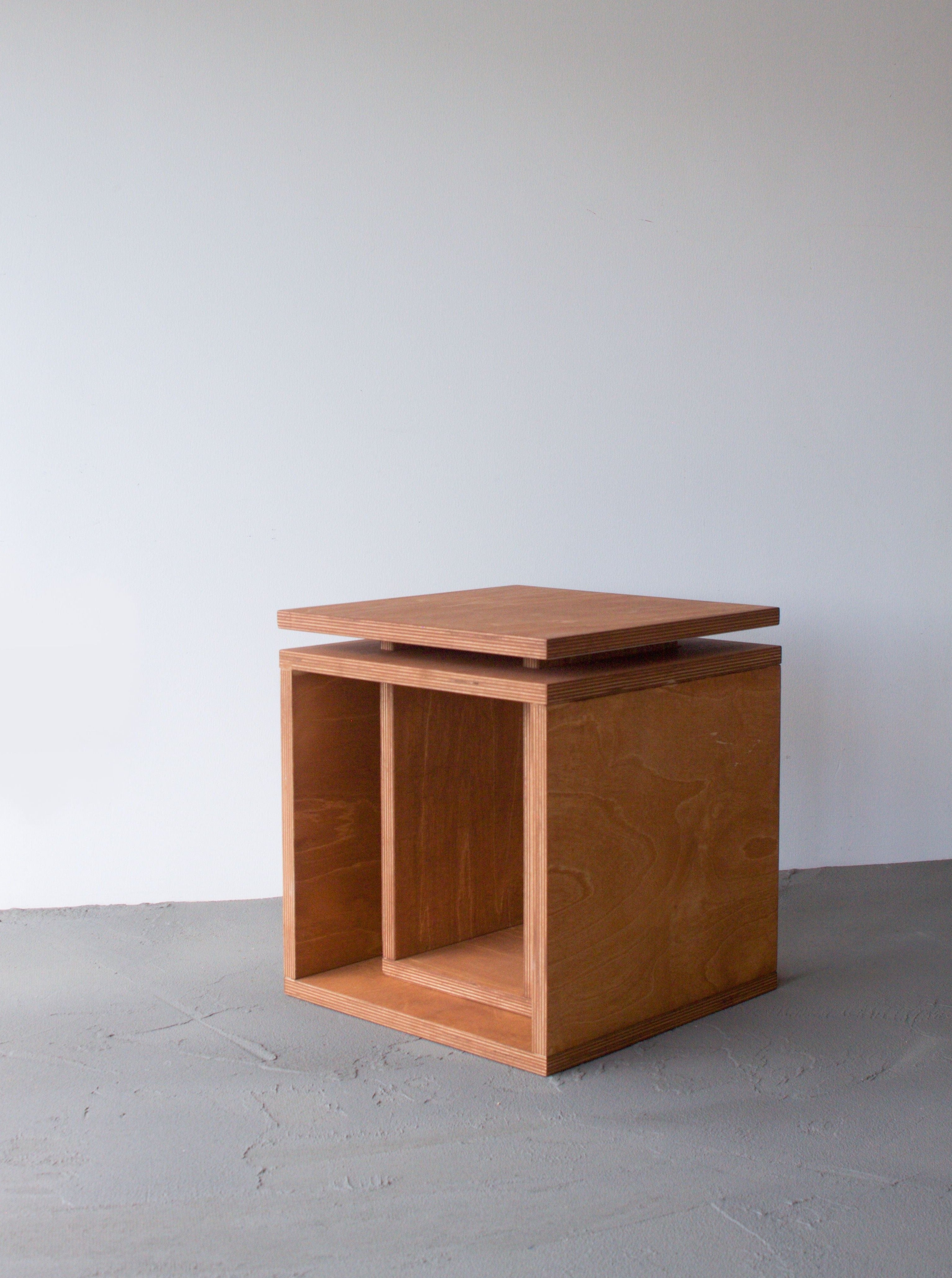 A minimalist Cubo side table from Edoardo Lietti Studio with an open shelf, positioned against a plain white wall on a textured gray floor.