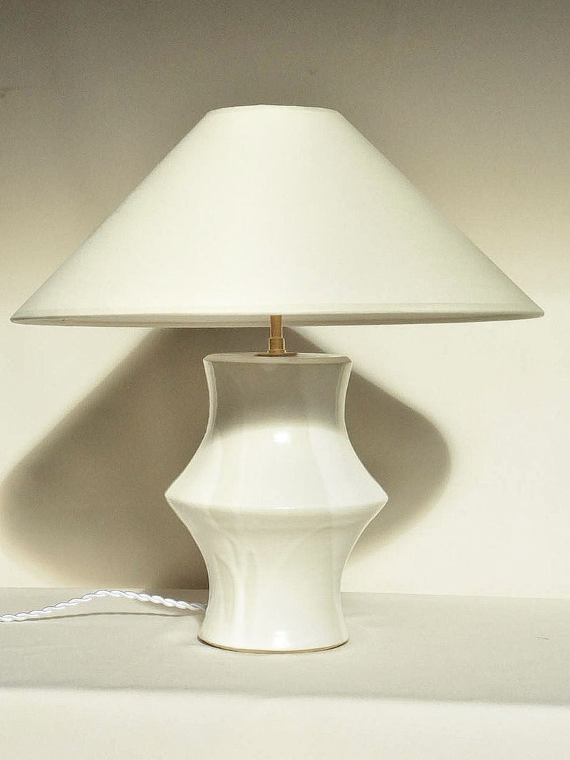 The 'Suzanne' Table Lamp by Landy Rakoto Ceramics features a handmade ceramic base with a wide, flared design and a simple cone-shaped shade. Set against a neutral background, its visible white cord adds Loire Valley-inspired charm.