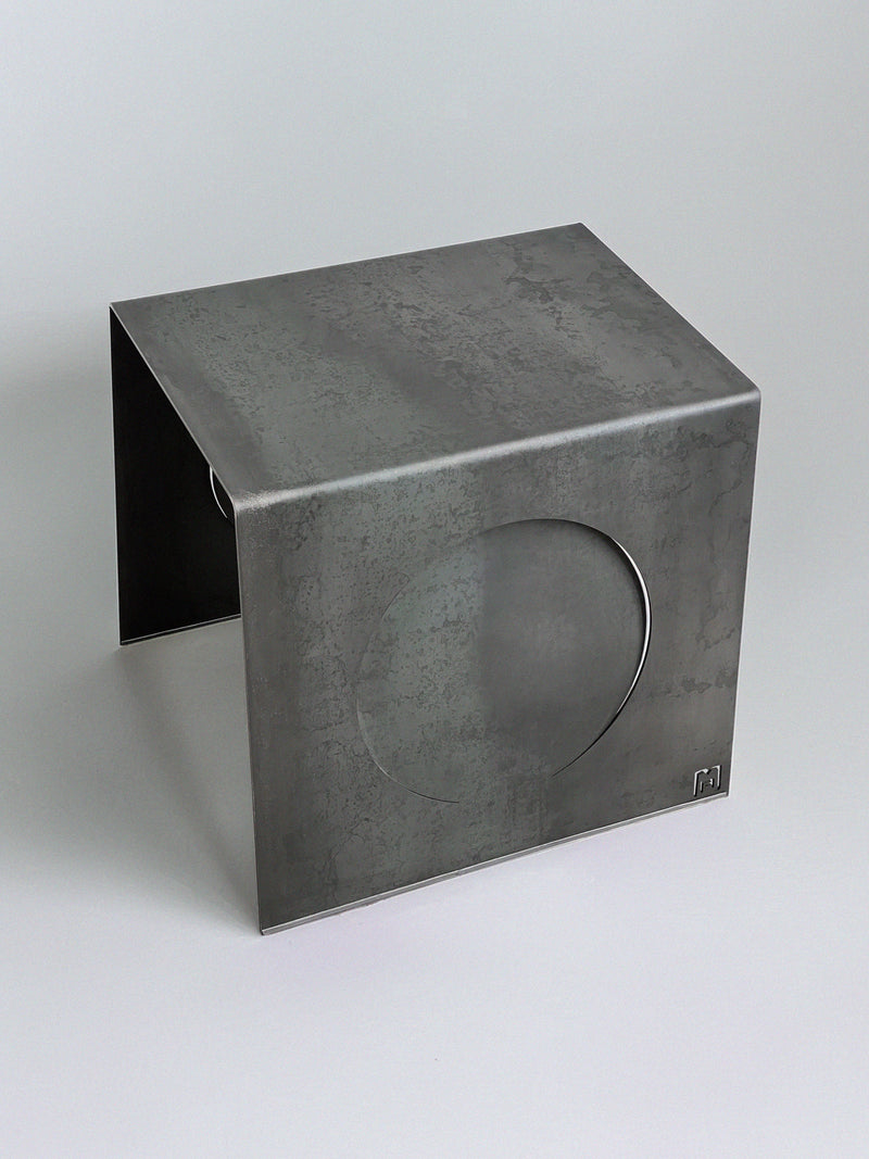The 'Era' Side Table by Modan features a minimalist, industrial design in matte gray steel with bent edges and a large circular cutout—subtle imperfections give each rectangular piece its own unique character.
