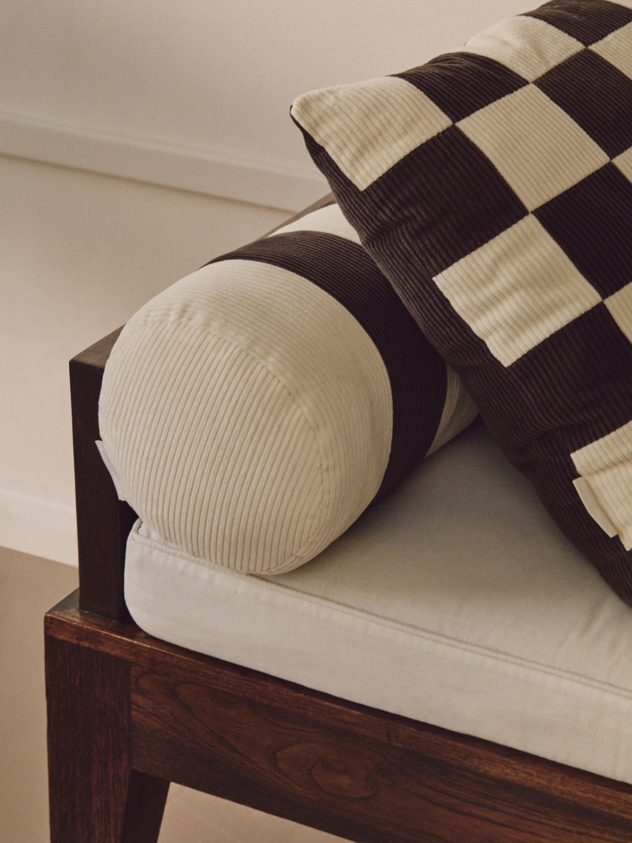 Close-up of a wooden-framed sofa with light-colored cushions and a cylindrical bolster pillow featuring a striped pattern. A large vintage-inspired patchwork cushion rests against the Le Cylindre - Cotton Corduroy - Espresso/Chalk by AMÉLIORÉ. The background is unfocused and neutral-toned, highlighting the handmade craftsmanship in the UK.