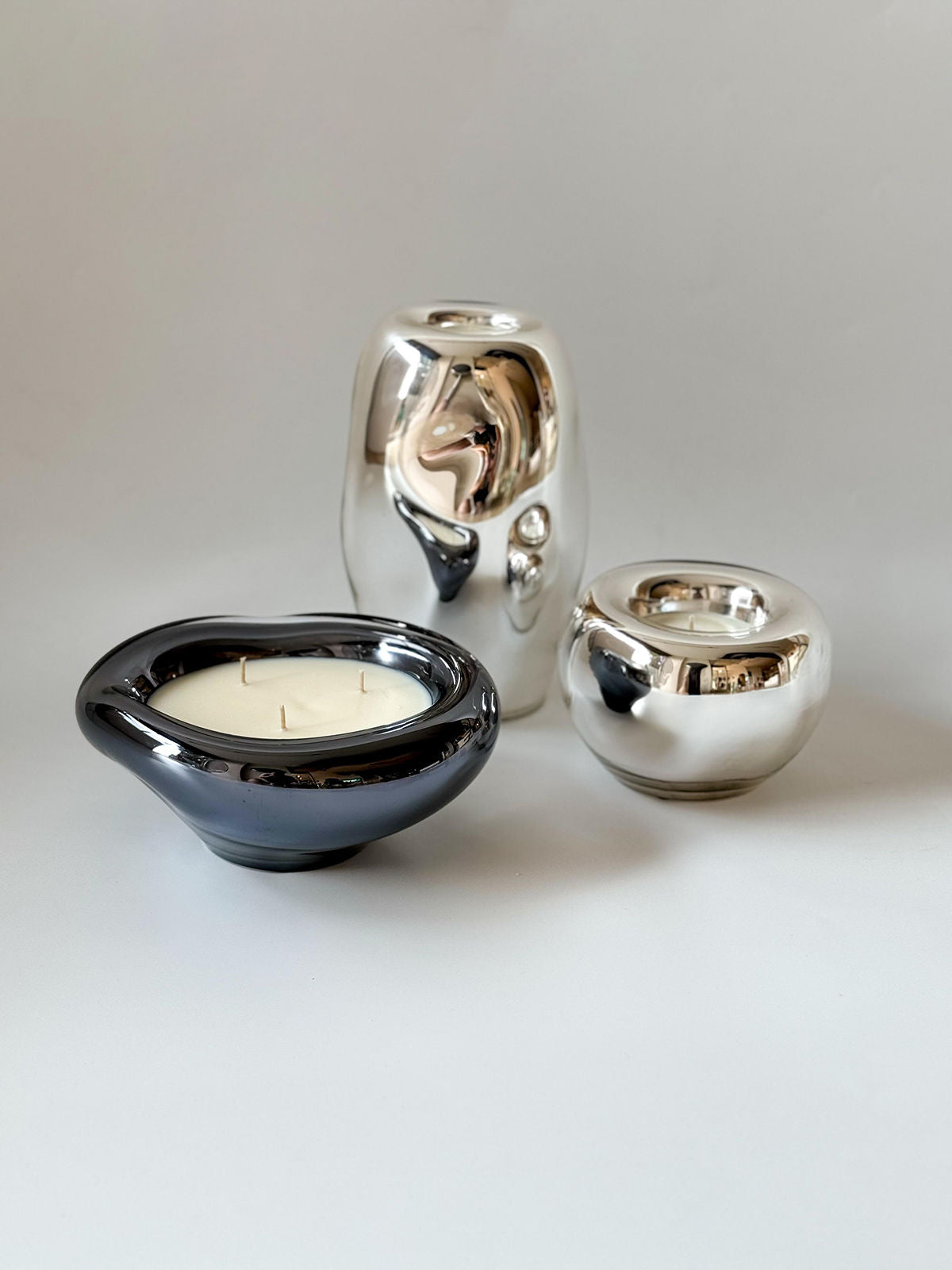 In the foreground, a black Lumni Candle by HOMA rests in a glossy, asymmetrical holder. Behind it, two reflective silver vases resembling elegant mirrors sit on a neutral white surface against a plain light gray background.