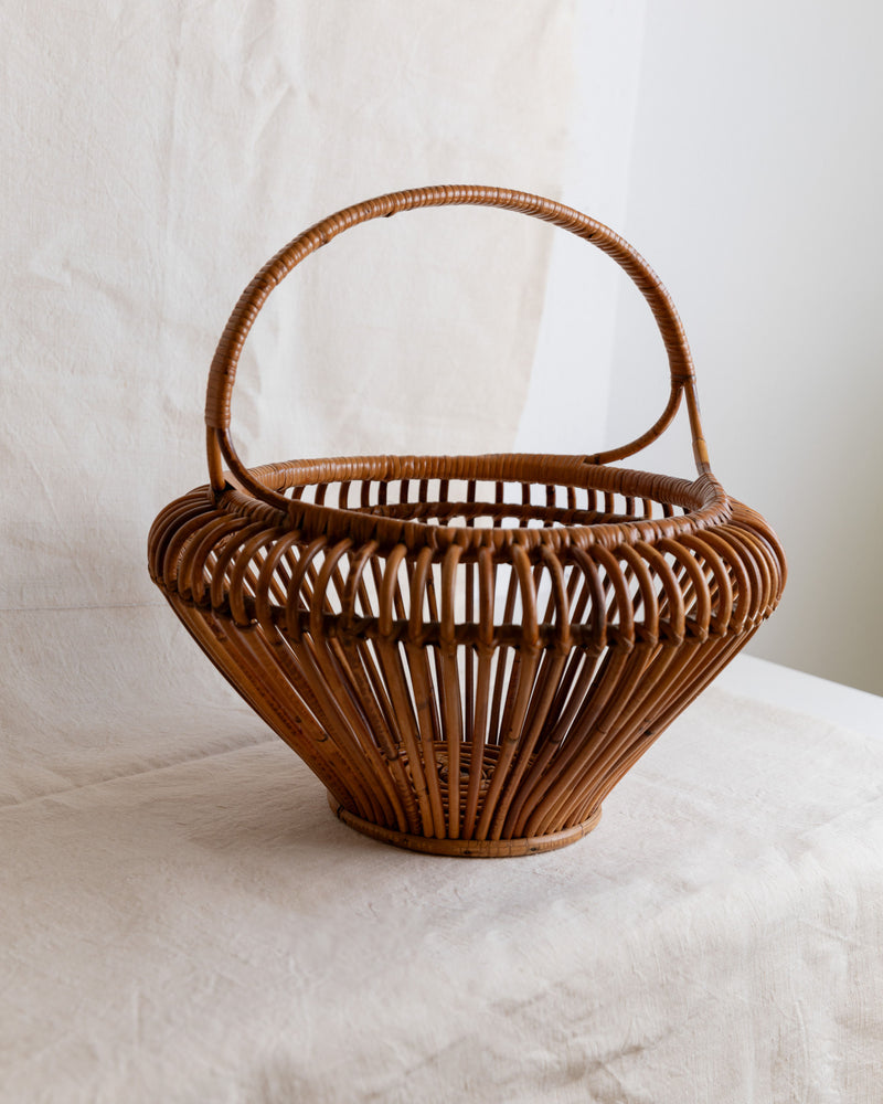 A round, open-weave wicker basket from Bottega Jacobs sits on a textured light surface. This XL Vintage Bamboo Basket Italy 1960s showcases mid-century flair with its rustic charm and delicate craftsmanship, emanating a minimalist aesthetic and casting subtle shadows.