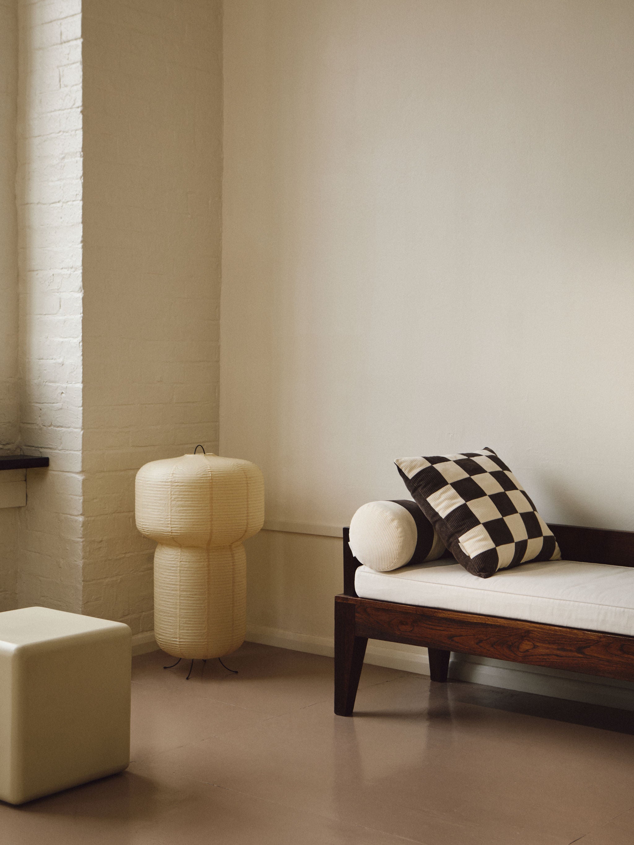 A minimalist living room with a white brick wall, a wooden bench featuring a white cushion and AMÉLIORÉ's Le Cylindre - Cotton Corduroy - Espresso/Chalk, a tall paper lantern beside the bench, and a small white ottoman in the foreground. The floor is light pink and the wall is beige.