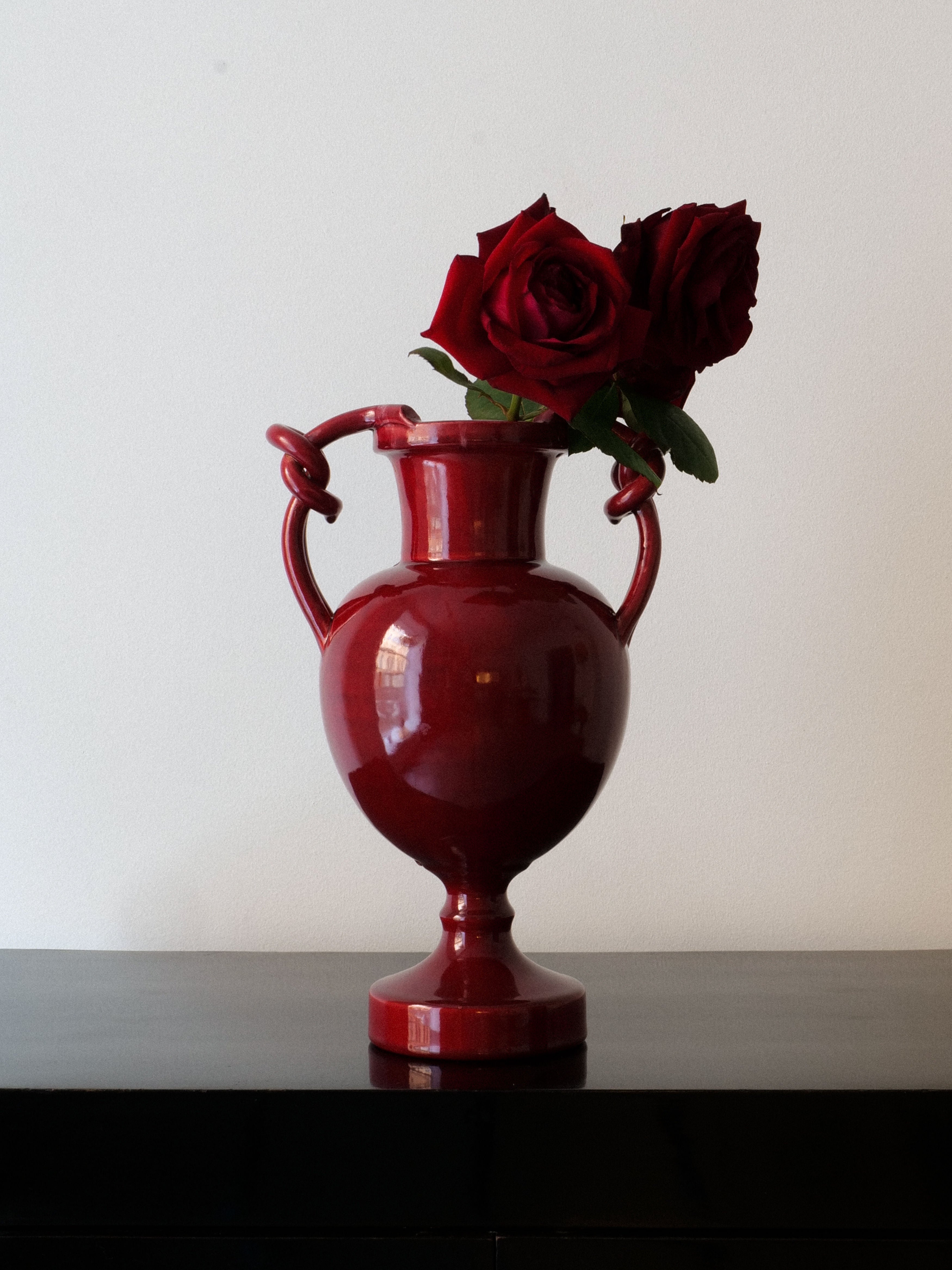 The Baluster Vase Jérôme Massier from Collection apart, featuring a classic design with two handles, evocative of the French Art Nouveau style, is displayed on a dark surface. It elegantly holds two vibrant red roses that create a striking contrast against the plain white background.