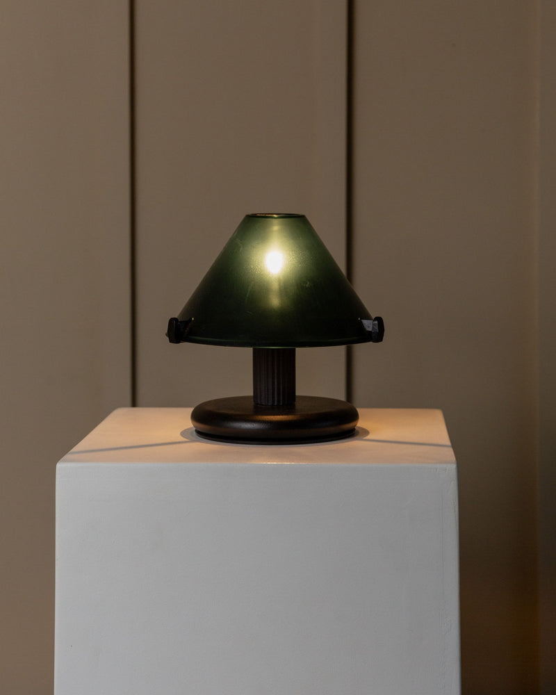 A small, dark green Vintage Table Lamp 80s from Bottega Jacobs, featuring a conical shade, is displayed on a white pedestal against a plain beige background. The decorative lighting is turned on, emitting a soft, warm glow.