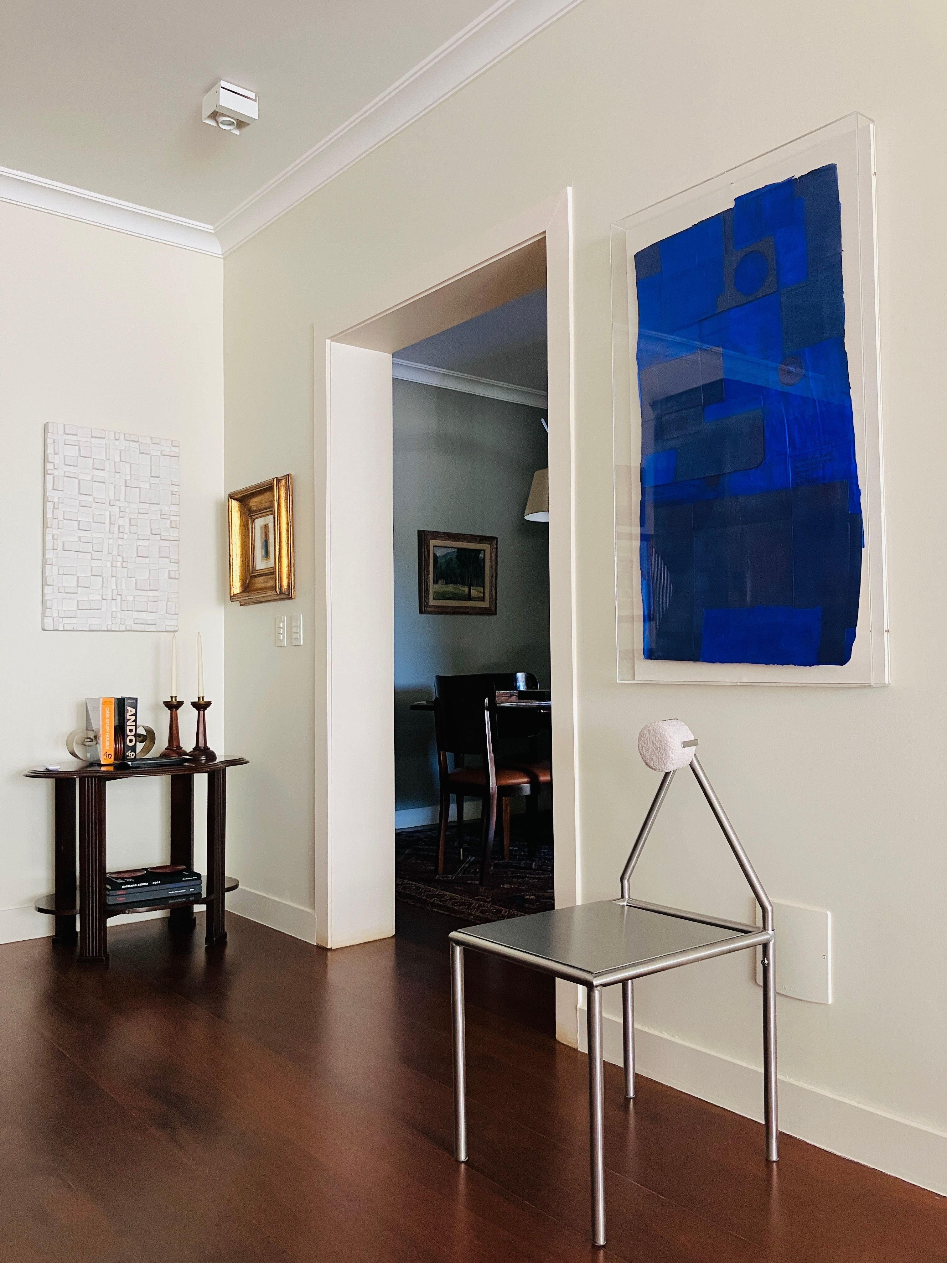 A modern living space with wooden floors, cream walls, minimalist furniture, abstract blue artwork, and the HT. 'H01' Stainless Steel Chair featuring a round white object on its seat. A small shelf displays candles and books.