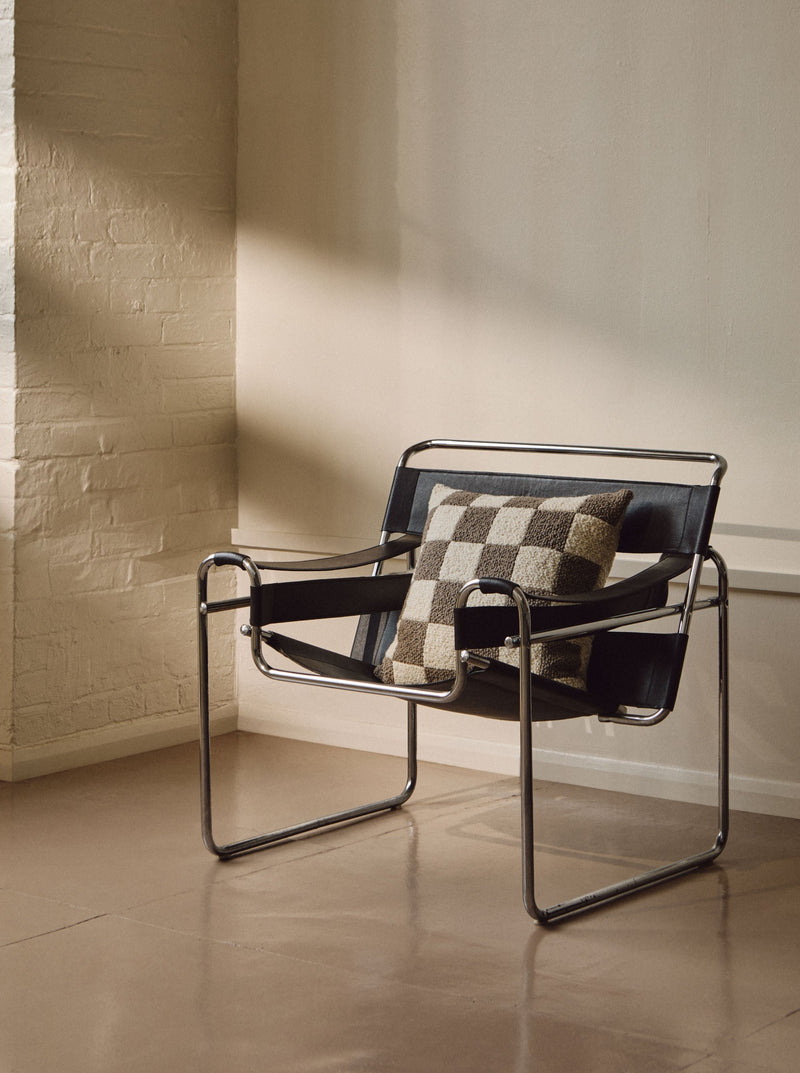 A modern armchair with a sleek chrome frame and black leather seating is positioned against a beige wall. The seat features a vintage-inspired design with a Le Carré - Wool Bouclé - Sawdust/Eggshell handmade cushion by AMÉLIORÉ. Soft natural light streams in from the left, creating a cozy atmosphere.