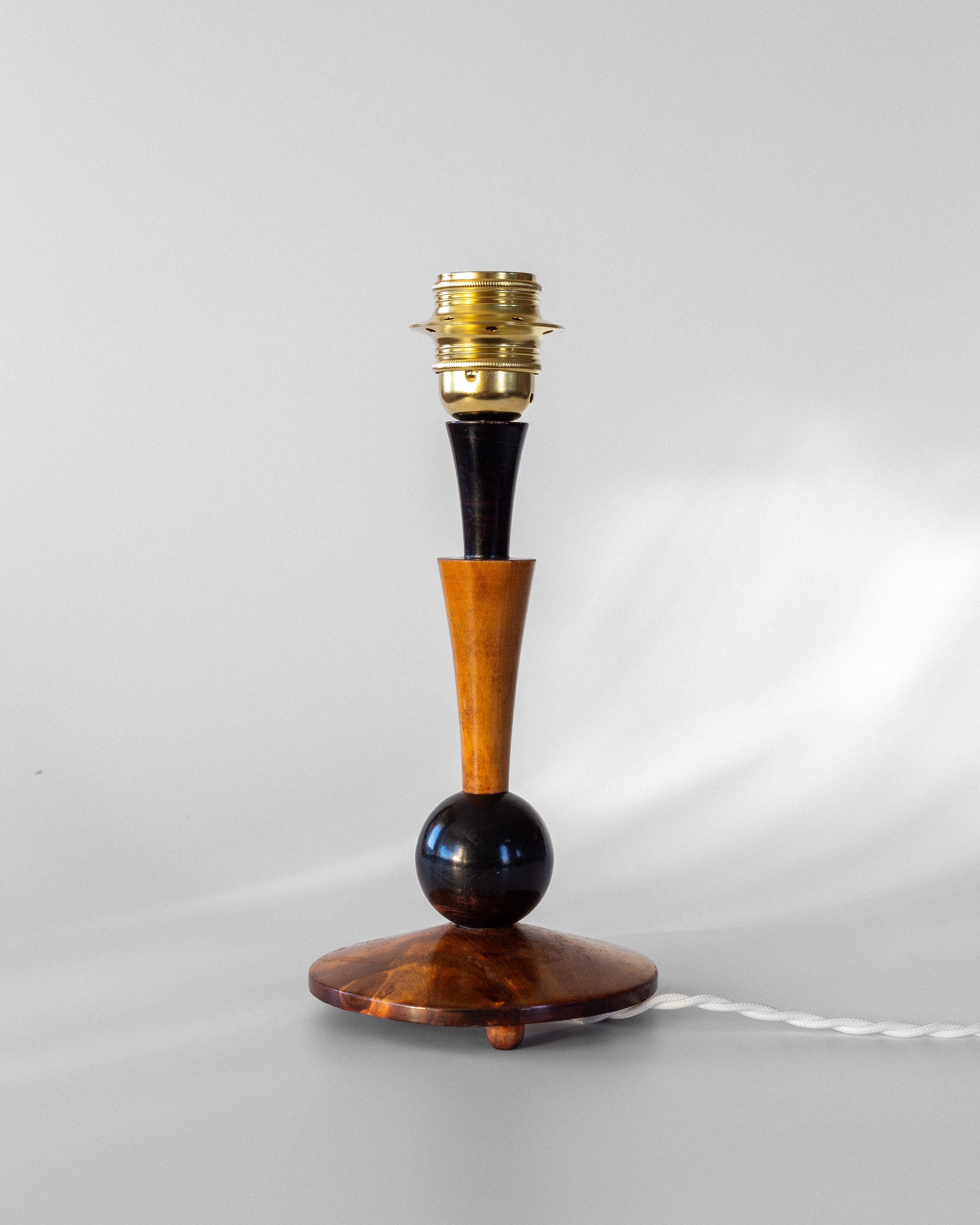 The Spigel Art Deco Wooden Table Lamp showcases a hand-crafted wooden base adorned with vintage character, featuring a dark, round central ball and tapering neck atop a flat, round base. Its brass socket harmonizes beautifully with the white cord. This exquisite piece radiates Art Deco charm against a plain light gray background.