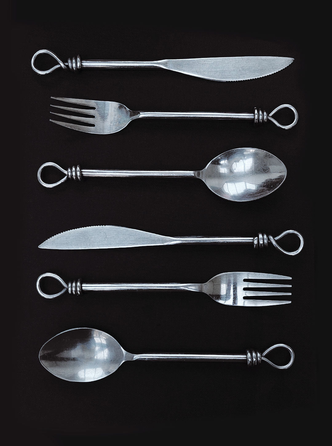 The Les Objoies Set of 6 Cutlery, featuring a stunning twisted loop design at the end of each handle, gleams beautifully against a black background. This sophisticated set includes two knives, two forks, and two spoons, ideal for enhancing your dining experience with an elegant touch.