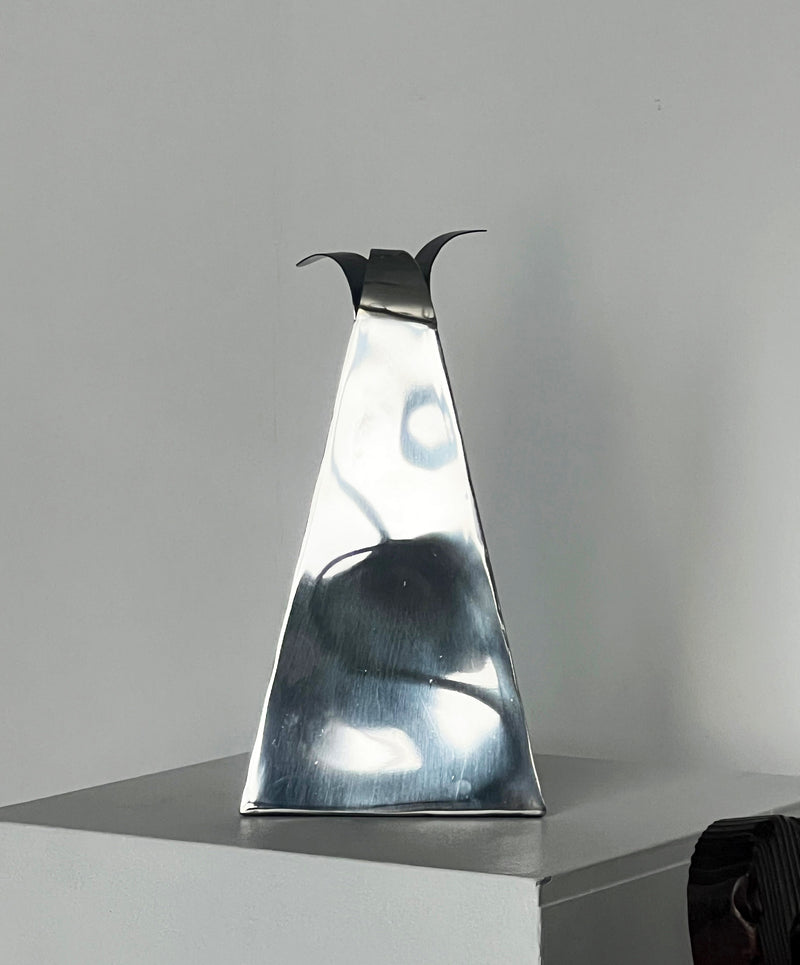 A reflective and stylish angular metal sculpture from Porter Studio, known as the Triangular Vase, features an elongated triangular form with two curved protrusions at the top. It is elegantly displayed on a pedestal against a plain wall.