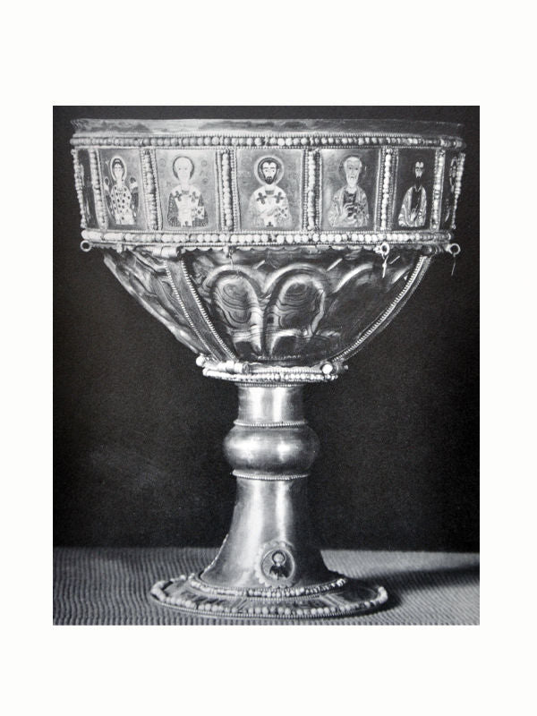 An ornate chalice made of metal with a prominent base. The middle section features intricately decorated panels with religious figures and symbols, framed by intricate beading. Reflecting Greek culture, the background is plain, highlighting the detailed craftsmanship of **The Greek World: Classical, Byzantine, and Modern** by **Maison Plage**.