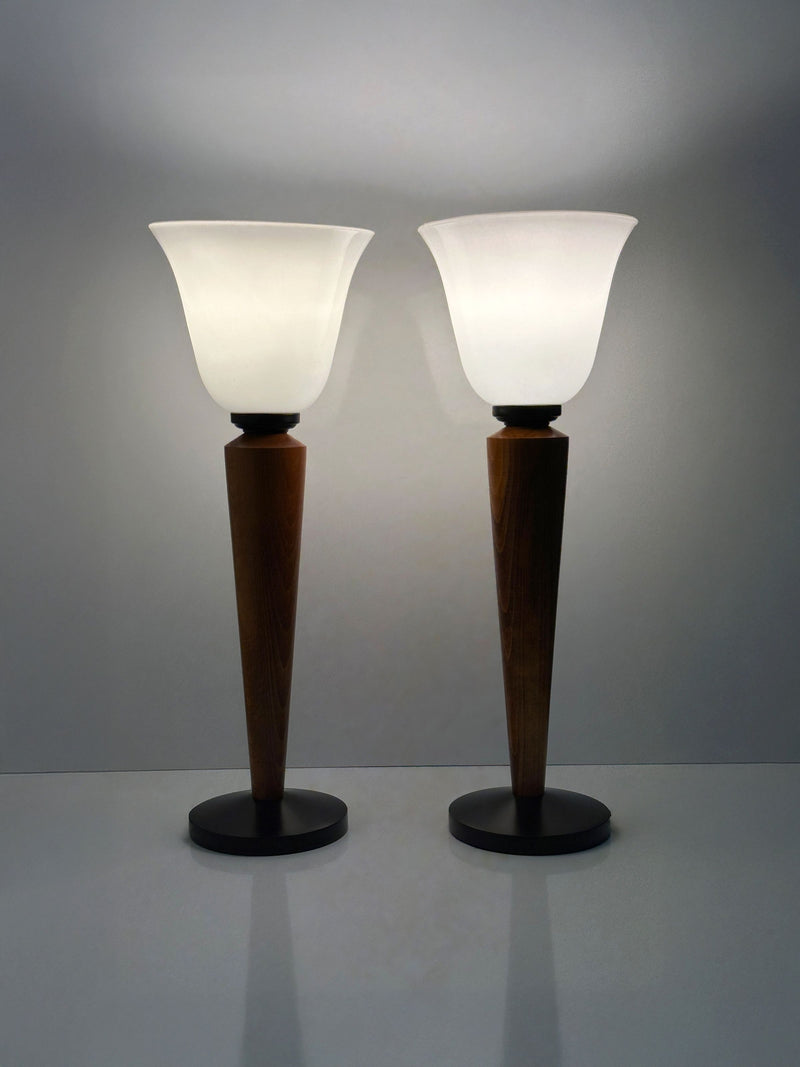 The Pair of Art Deco Chalices with Teak Wood by Dodo Vintage stand side by side on a smooth surface against a plain background, featuring frosted glass shades and teak stems. They emit a soft, white glow, casting gentle reflections below.