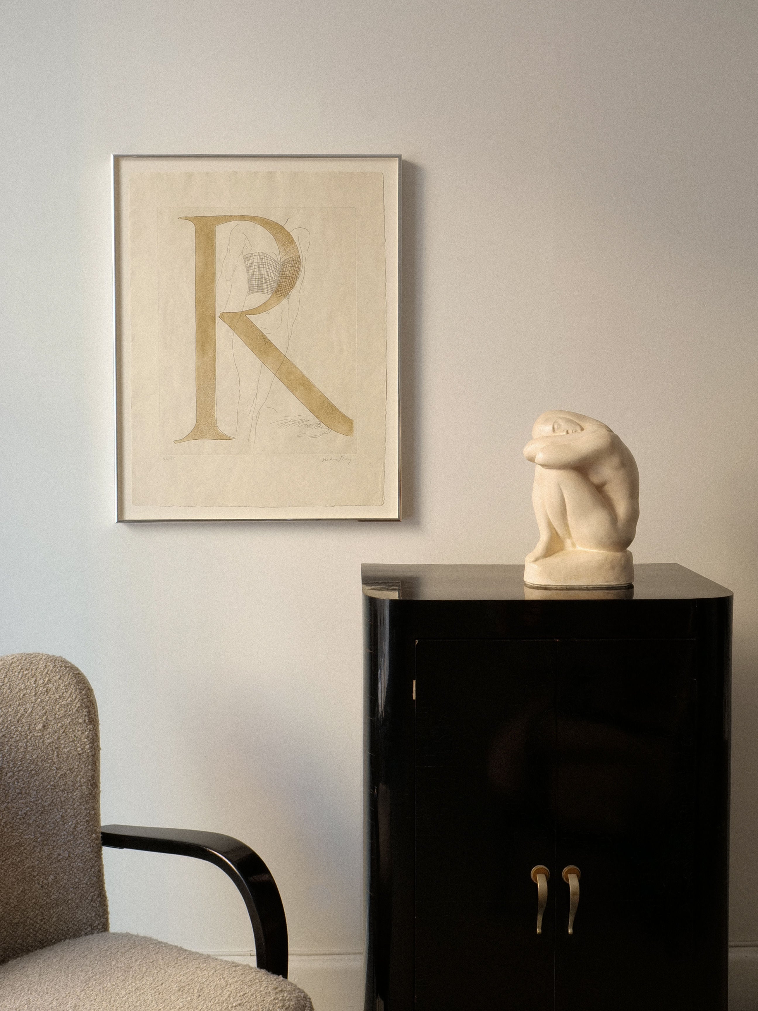 In a minimalist room, the focal point is a framed letter "R" artwork on the white wall. Nearby, a black cabinet displays Collection Apart's Resting Sculpture by Bror Forslund, complementing the textured gray chair with its black armrest visible on the left.