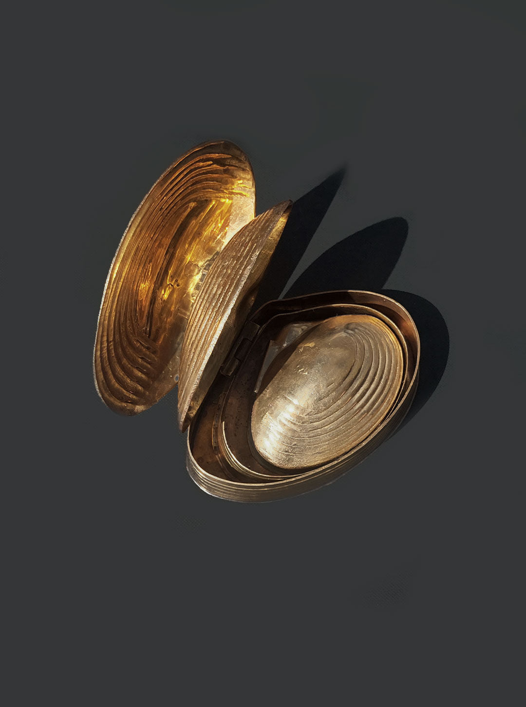 A Set of 3 Nesting Shell Boxes by Les Objoies with a gold finish shaped like a clamshell, slightly open to reveal its reflective inner surfaces. The metallic object, adorned with shell patterns, is photographed against a dark background, casting shadows beneath it.