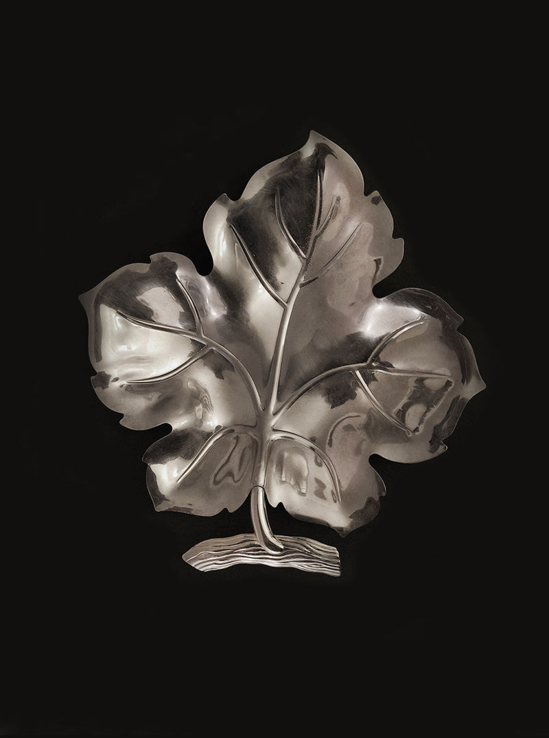 Against a plain black background, the Wine Leaf Vide-Poche by Les Objoies is a metallic sculpture with five lobes, embodying minimalist design. Crafted from stainless steel, it features a textured stem and smooth surfaces.