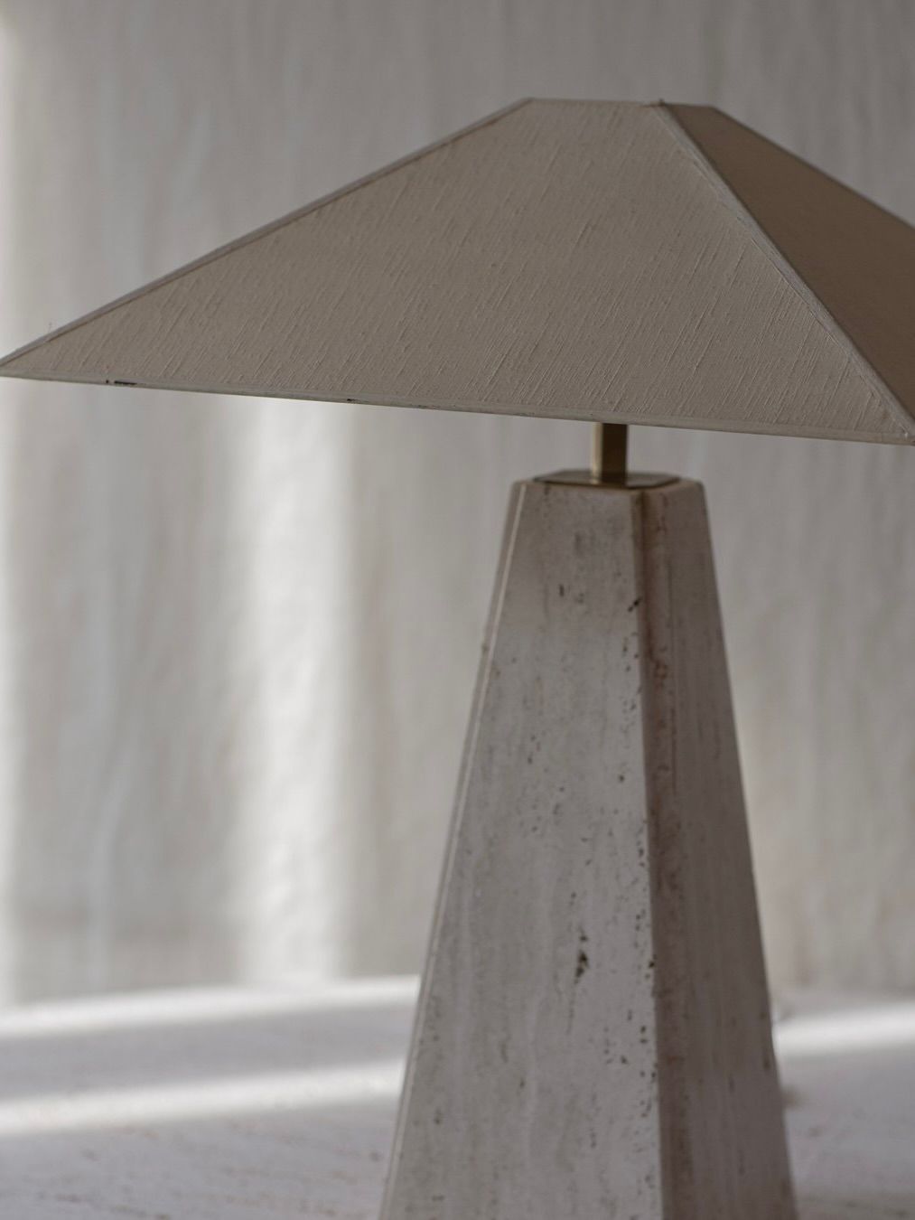 The Bicci de' Medici Camille Breesch - Travertine Table Lamp features a minimalist, cement-textured pyramid base and wide triangular shade. Its elegant geometric form stands out on the light surface against a softly blurred background.