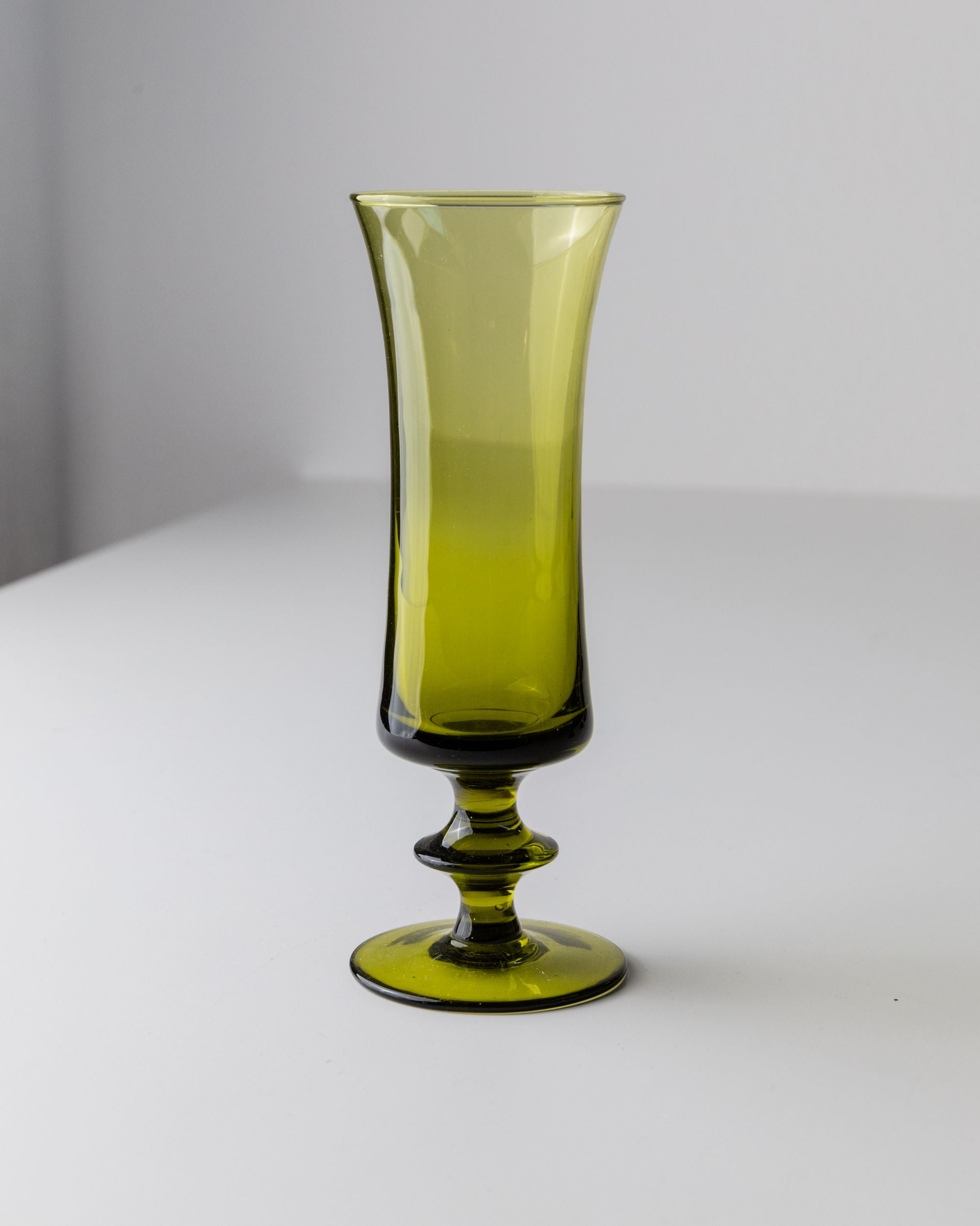 A tall, elegant glass from Bottega Jacobs' 4 Murano Glasses in Green 70s set stands on a white surface, featuring a flared rim and decorative stem. It casts a soft shadow to the right on a neutral wall, epitomizing retro chic style with mid-century design.