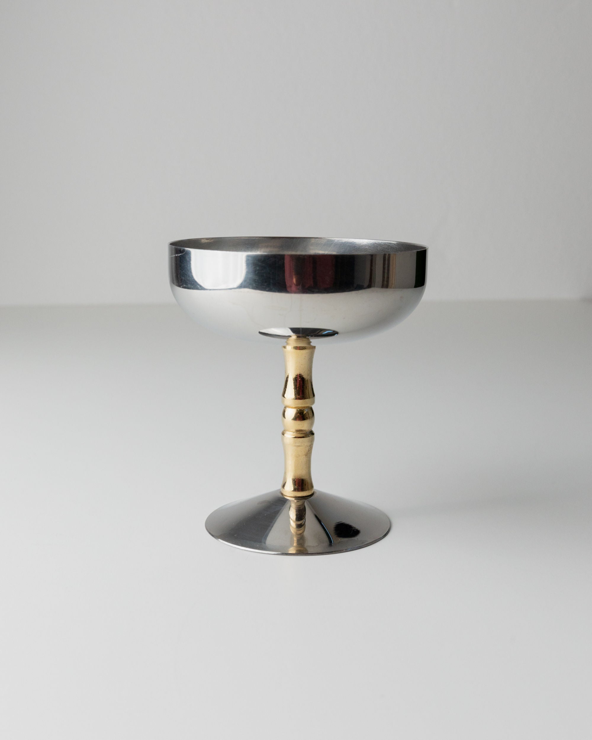 A Bottega Jacobs "6 Stainless Steel Champagne Coupes 70s" goblet, with a wide shallow bowl and metallic stem resembling silver and gold vintage stainless steel, sits on a plain white surface against a light grey background.