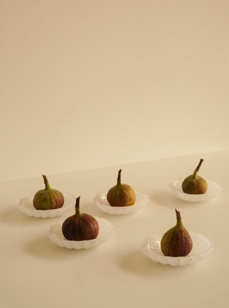 Five ripe figs are placed individually on delicately crafted **5x Shell Dessert Plates** by **Les Objets Amsterdam** in a white, scalloped design on a light-colored surface with a cream background. The arrangement is evenly spaced, giving a minimalist and elegant appearance.