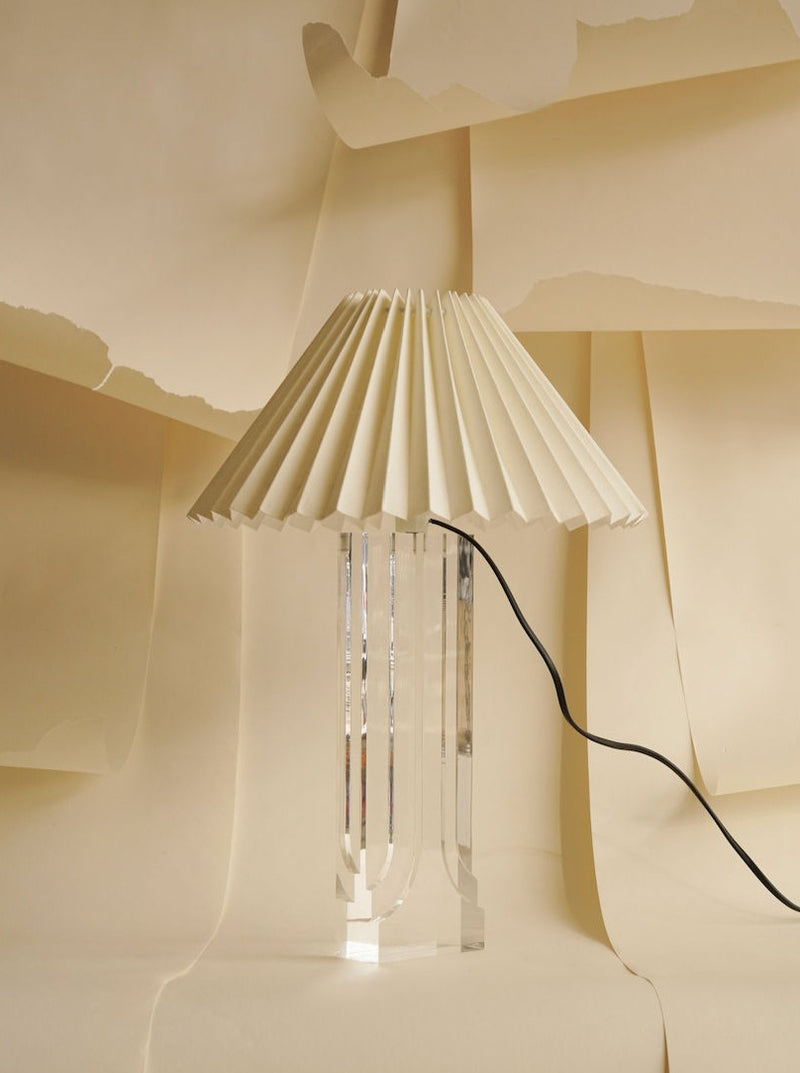 An Art Deco "Ghost" Lamp by Les Objets Amsterdam stands against a beige backdrop of layered and torn paper. The classic lamp features a pleated, off-white lampshade and a visible black power cord. The setup creates a minimalistic and elegant visual effect.