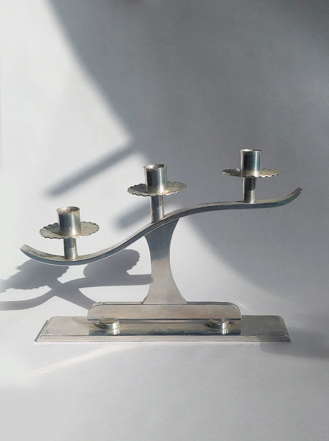 A pair of metallic candle holders by Les Objoies, known as the "Pair of Wavy Candle Holders," features a modern design with three candle holders each mounted on wavy, elevated bases. These pieces cast shadows on a light background and boast symmetrical structures with minimalist forms. Geometric accents further enhance their contemporary aesthetic.